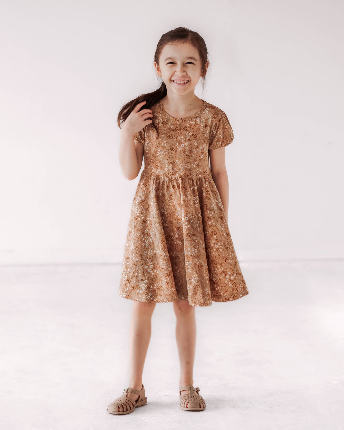A young child wearing a brown floral twirl dress with puff sleeves and a wide skirt.