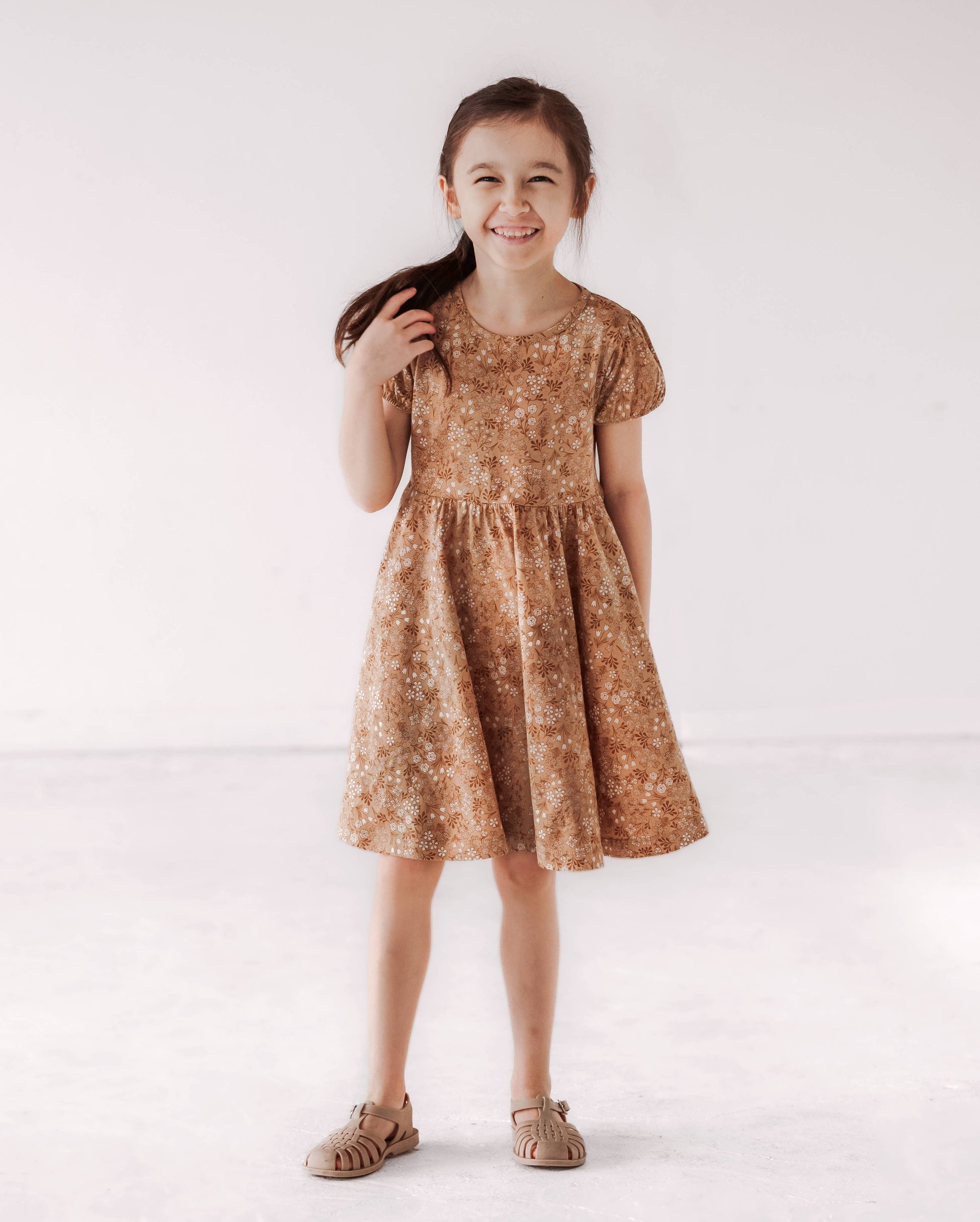 A young child wearing a brown floral twirl dress with puff sleeves and a wide skirt.